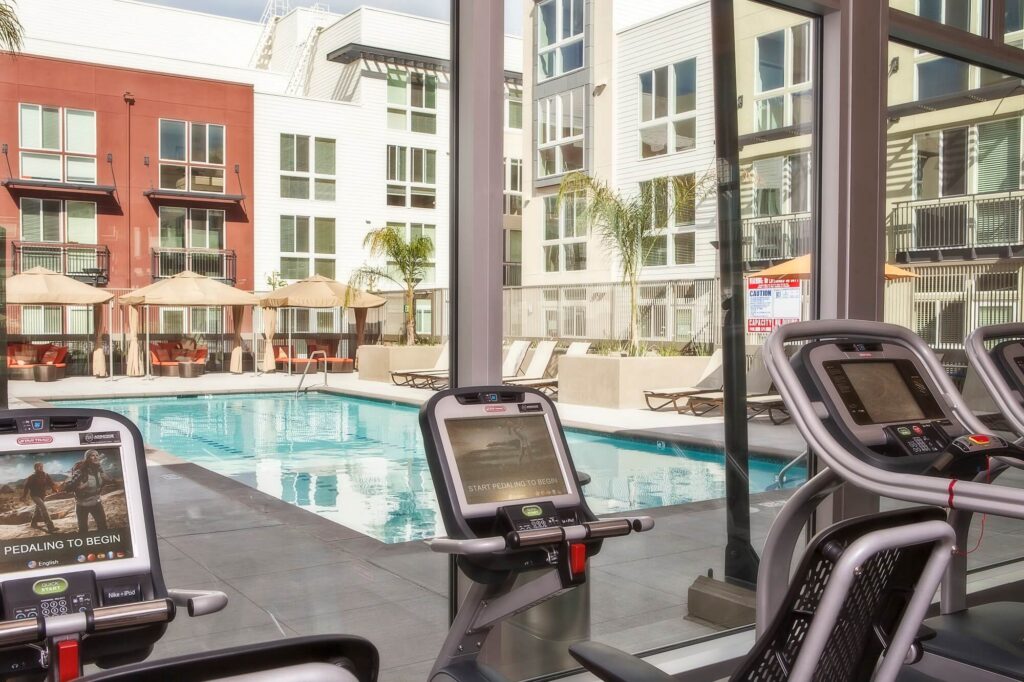 Modern gym with exercise bikes overlooks an outdoor pool with lounge chairs, umbrellas, and surrounding apartment buildings through large floor-to-ceiling windows.