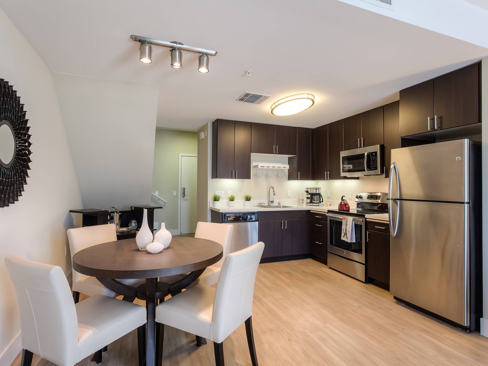 Modern kitchen and dining area with dark cabinets, stainless steel appliances, a round table with four white chairs, and decorative vases. Bright lighting and wooden flooring complete the contemporary look.