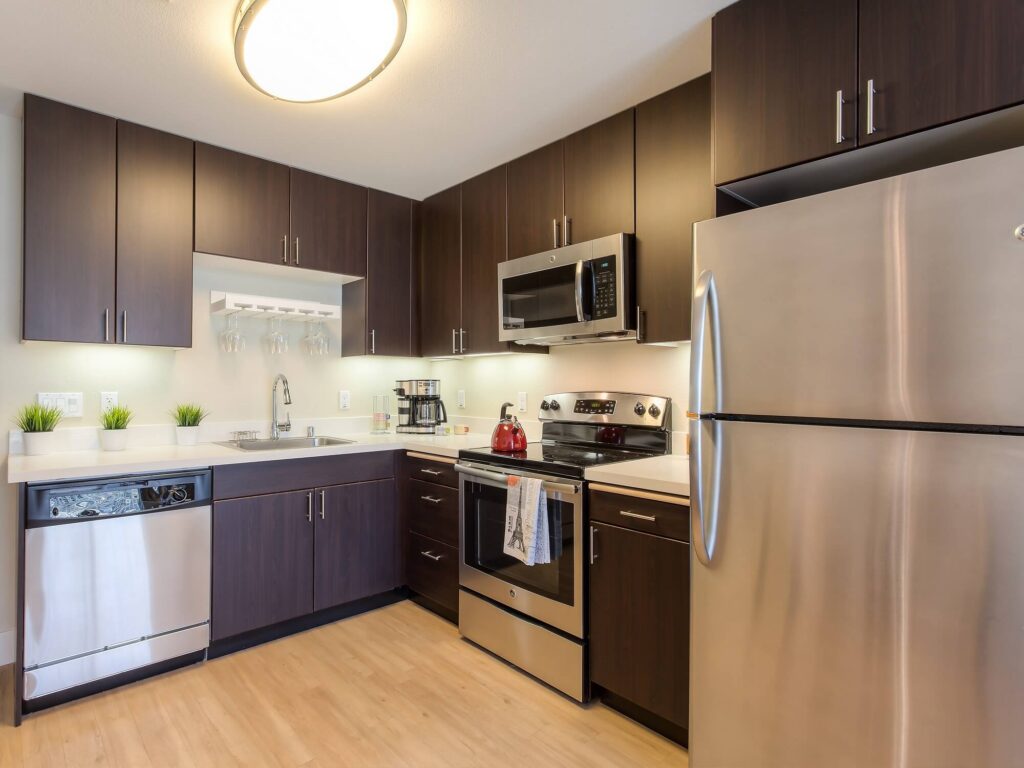 Modern kitchen with dark wood cabinets, stainless steel appliances, light wood flooring, small potted plants, a dishwasher, stove with a kettle, microwave, and fridge. Countertops are white and well-lit by overhead lights.