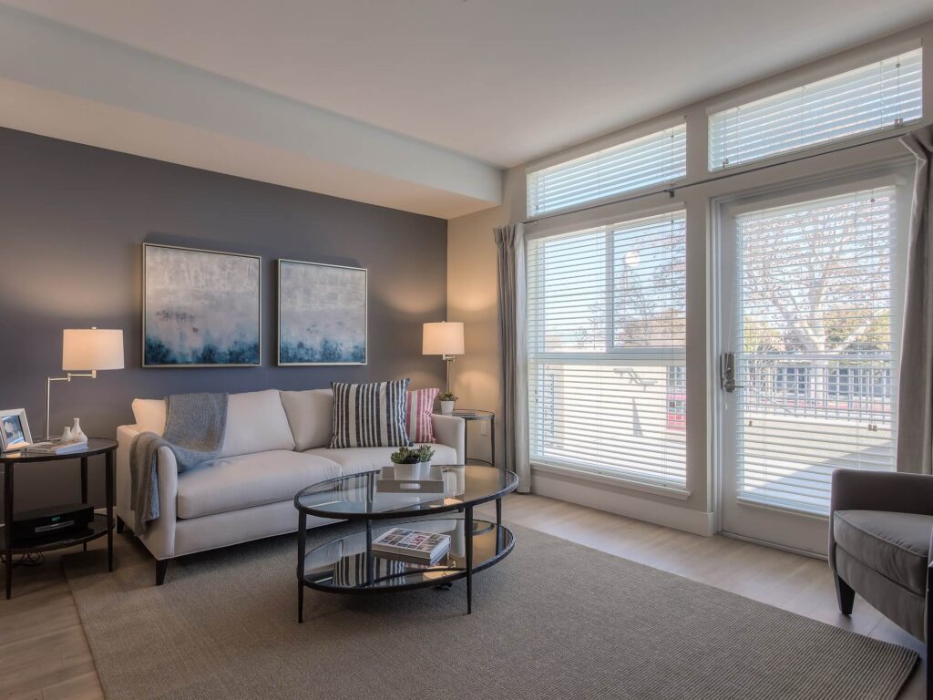 A modern living room with a beige sofa, glass coffee table, two lamps, abstract art on a gray accent wall, large windows with blinds, and a balcony view.