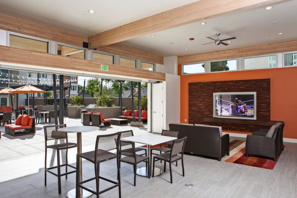 Modern lounge area with tables, chairs, and sofas facing a wall-mounted TV; large glass doors open to a patio with outdoor seating and string lights.