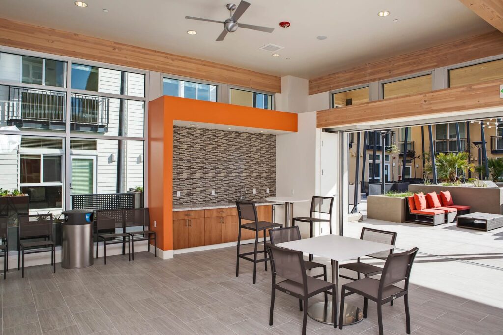 Modern indoor lounge with tables, chairs, and a small kitchen area. Large windows and sliding doors open to an outdoor patio with couches. Light wood and orange accents create a bright, welcoming space.