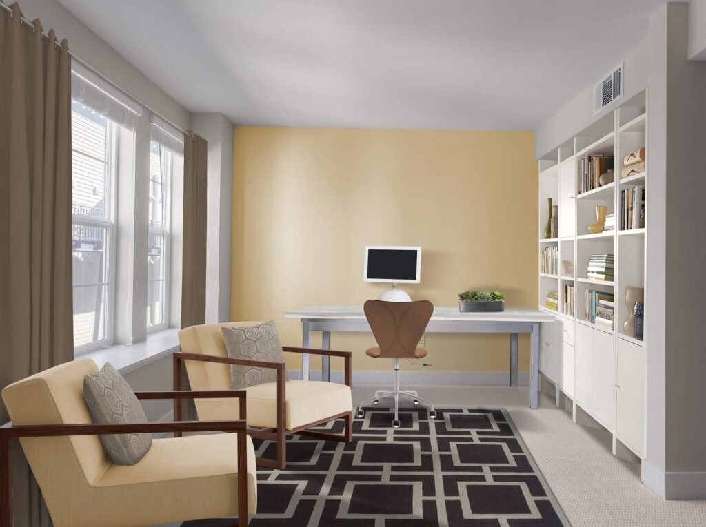 Bright home office with a desk, computer monitor, modern chair, bookshelves, two beige armchairs, patterned rug, large window with curtains, and a potted plant on the desk.