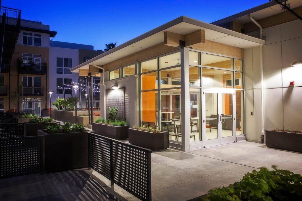 Modern building exterior at dusk, featuring large glass windows, illuminated interior, surrounding planters, and an outdoor patio area with seating. The sky has a blue evening glow.