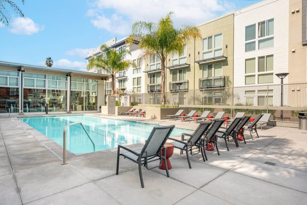 Modern apartment complex with a rectangular outdoor pool, multiple lounge chairs, palm trees, and large windows. The sky is clear with a few clouds, and the pool area looks clean and inviting.