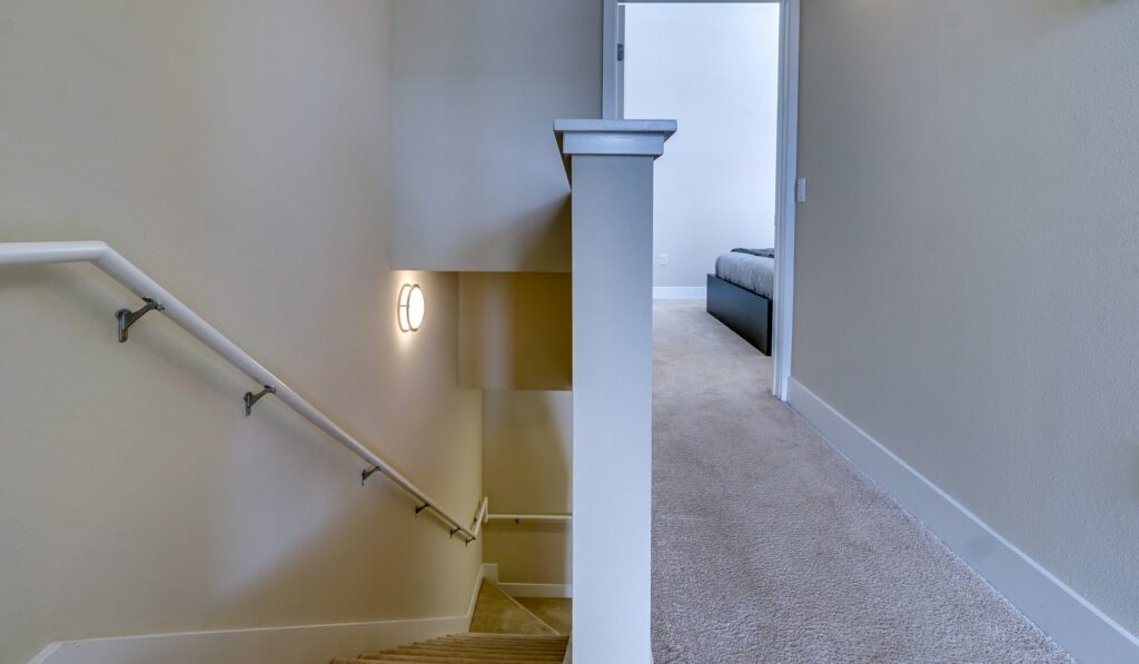 A beige-carpeted hallway leads to a bedroom. Stairs with white railings descend to the left, and a wall-mounted light illuminates the stairwell. The walls and trim are light-colored.